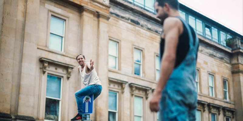 Person balancing on top of a street sign while another stands in the foreground in front of a historic stone building.