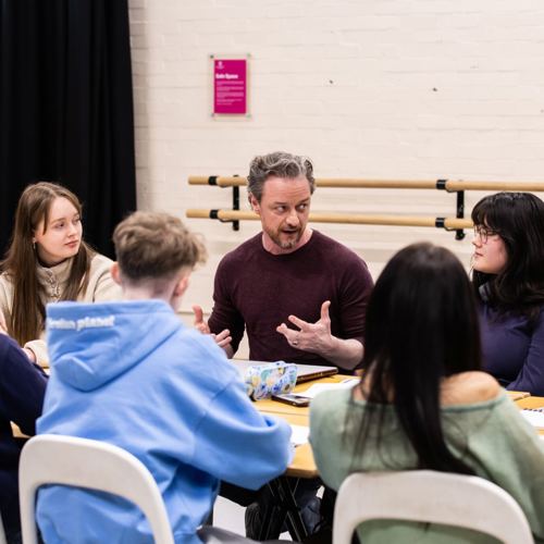 A group of people, seated around a table, engaged in a lively discussion. The setting is casual, with notebooks visible. The mood is focused and collaborative.