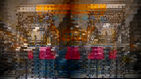 The opulent bar area of Anchor Line restaurant, featuring a well-stocked back bar with bottles and glasses, red bar stools, and elegant decor.