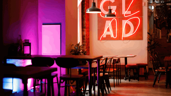 Modern café interior with wooden tables, pink and purple lighting, hanging lamps, neon “GLAD” sign, and greenery creating a cozy, stylish atmosphere.