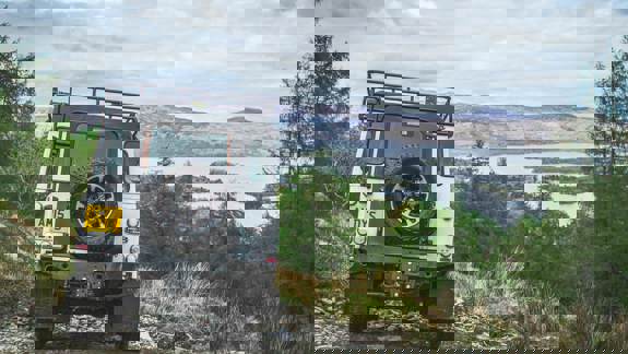 A white Land Rover on a rugged off-road trail overlooking Loch Lomond and surrounding hills.