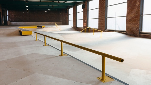 An empty indoor skate park with wooden ramps and a long yellow rail. The space is a converted warehouse with brick walls and large, frosted windows.