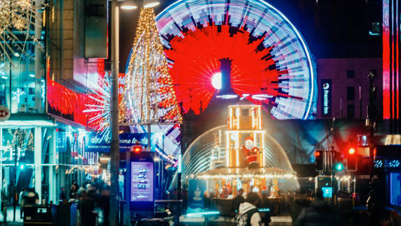 Night scene of a Ferris wheel glowing intense red and white, a large illuminated Christmas tree, and a festive Santa's Grotto dome in a busy city square.