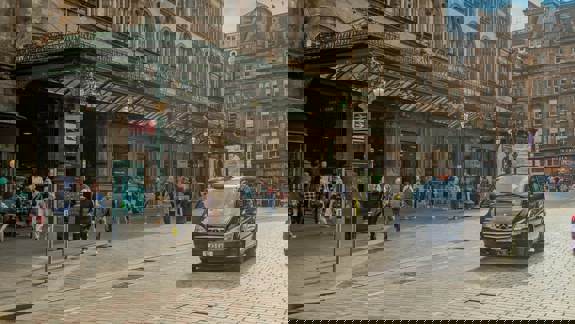 People walk past the historic entrance of Glasgow Central Station, while a black taxi drives along the cobbled street. 