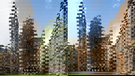 University of Glasgow main building with Gothic spire and cloisters, viewed across a green courtyard.