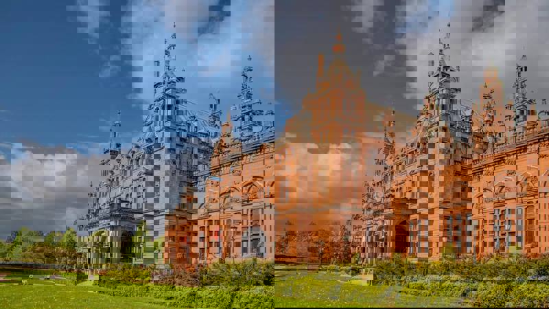 A large, ornate red sandstone building with multiple towers and a curved entrance is surrounded by a green lawn and trees under a blue sky.