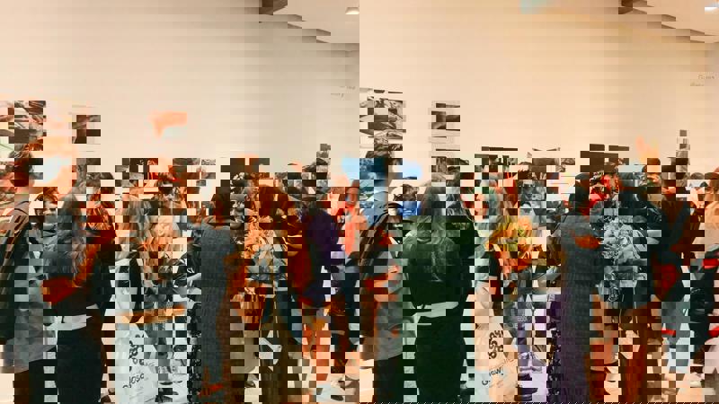 A crowded art gallery opening with a diverse group of people viewing photographs and installations on white walls. In the foreground, a woman holds a bouquet of flowers.