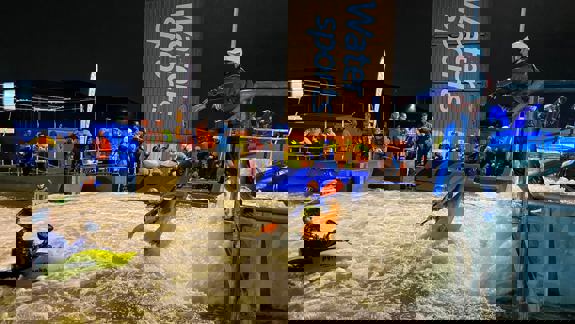 Kayakers in helmets tackle white-water rapids at Pinkston Watersports, with spectators and instructors watching from the side.