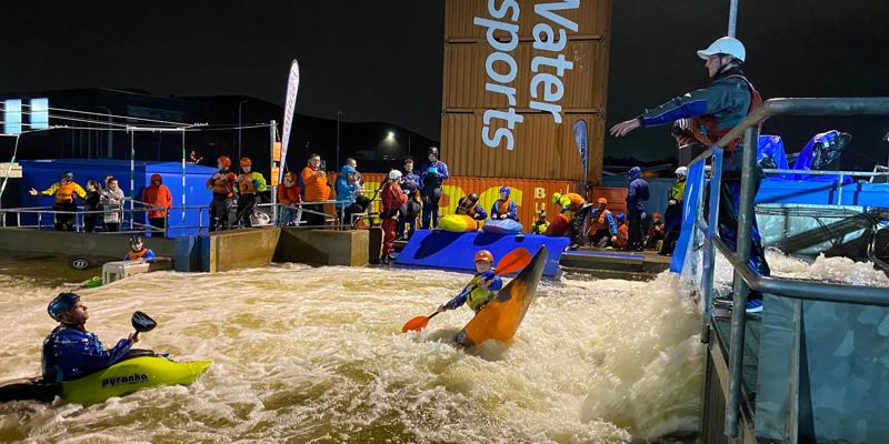 Kayakers in helmets tackle white-water rapids at Pinkston Watersports, with spectators and instructors watching from the side.
