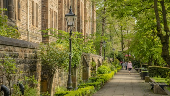 Old style lampposts, green foliage and trees line a pathway beside a historic stone building on the University of Glasgow campus.