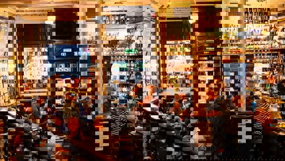 Traditional pub interior with warm lighting, groups chatting over drinks, and a bartender serving at a liquor-stocked bar. Vintage décor and ornate details create a cosy atmosphere.