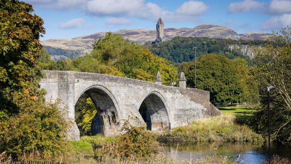  A historic stone bridge with two arches crosses a river. In the background, the Wallace Monument stands tall on a hill with a backdrop of a clear sky and clouds.