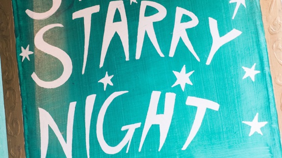 A close-up of a rustic, hand-painted sign on a green background with a gold frame that reads "STARRY STARRY NIGHT" in white, capital letters surrounded by stars.
