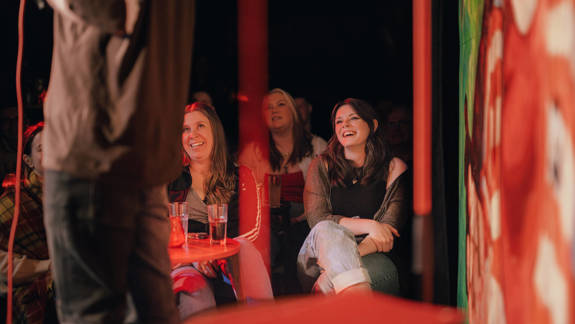 Audience members laugh during a performance at a comedy club. The shot is framed from behind the performer, focusing on the joyful reactions in the crowd.