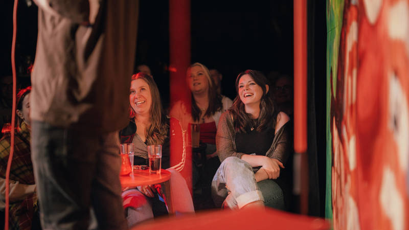 Audience members laugh during a performance at a comedy club. The shot is framed from behind the performer, focusing on the joyful reactions in the crowd.