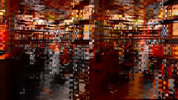 A dark, cosy bar and bookstore with wood-panelled walls and a patterned ceiling. Bookshelves filled with books surround the room. People sit at tables, enjoying drinks.