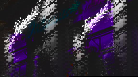 An exterior shot of a building at night, with a bright purple neon sign that reads 'Delmonica's' shining above the entrance.