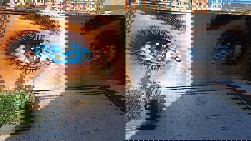 Close-up mural of large, vivid blue eyes on a building wall next to a paved area with benches.