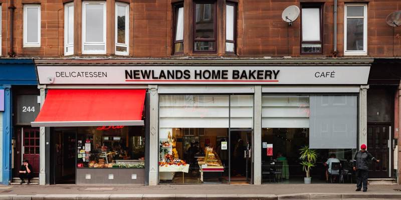 A storefront view of Newlands Home Bakery and Cafe in Glasgow, featuring a red awning and window displays of baked goods.