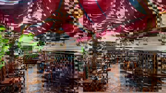 Balcony in the Polo lounge with wooden floors, tables and chairs as well as red fabric hanging from the ceiling.