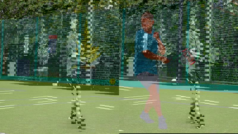 A male tennis player in a teal shirt and black shorts is in mid-swing on a green outdoor tennis court. A tennis ball is in mid-air in front of him.