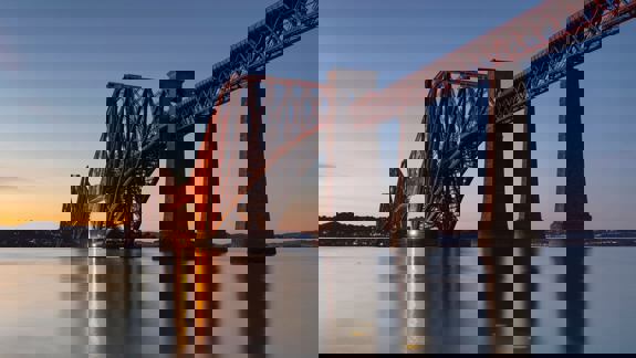 The iconic red Forth Bridge, a cantilever railway bridge, stretches over calm water at dusk, its lights glowing.