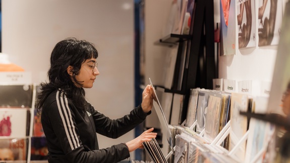 A young person with dark hair and glasses in a black tracksuit jacket is browsing vinyl records in a brightly lit record store.