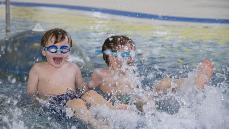 Two young boys with swimming goggles are splashing and laughing in a pool, creating a large splash of water. One boy is looking directly at the camera.