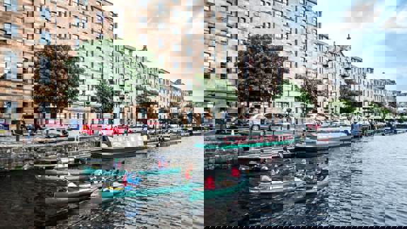 Groups of people in canoes paddle along the canal in front of historic stone tenement buildings in Glasgow.