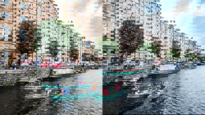 Groups of people in canoes paddle along the canal in front of historic stone tenement buildings in Glasgow.