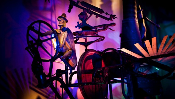 A kinetic sculpture of a skeleton in a top hat on a unicycle in a dark room with colourful lighting.