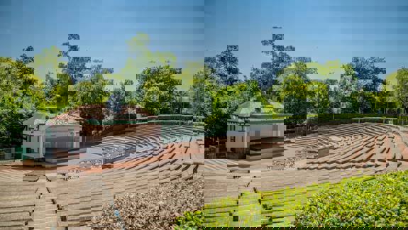 An empty outdoor amphitheatre with concrete benches and a central bandstand, surrounded by green trees under a clear blue sky.