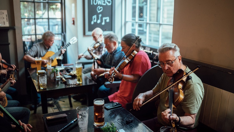 A group of people play traditional music in a pub. The musicians are seated at a table with pints of beer, playing fiddles and a guitar. 