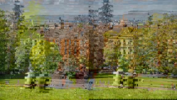 Two people sit on a grassy hill in Queen's Park, with a panoramic view of the Glasgow skyline and distant hills behind them.