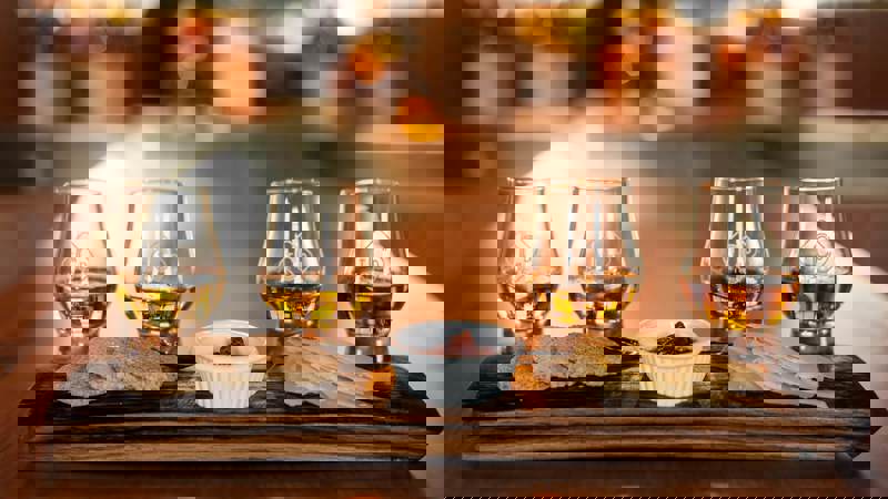 Four glasses of amber whisky are arranged on a wooden serving board with cheese and crackers.