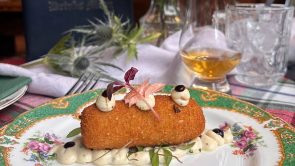 A golden-brown, breaded croquette is plated on a decorative floral plate, resting on a base of piped creamy sauce. The croquette is topped with small dollops of sauce, dark glaze, and delicate pink garnish with microgreens. In the background, a table setting includes a glass of amber-colored drink, water glasses, cutlery, and soft floral décor, creating an elegant dining atmosphere.