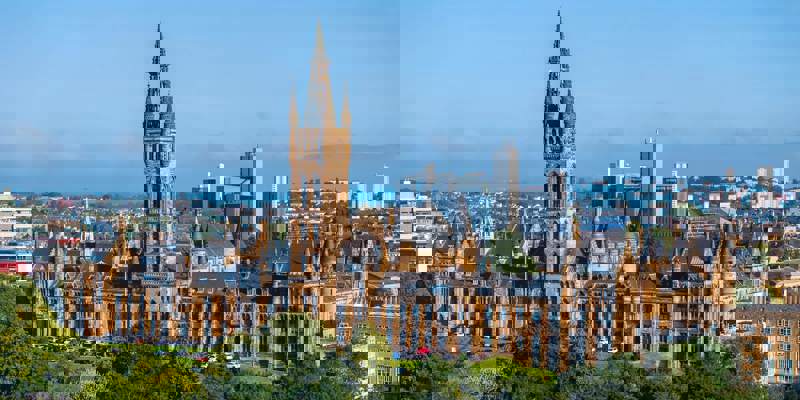 A panoramic shot of the historic Glasgow University building with its striking Gothic revival architecture and prominent clock tower, set against a blue sky with the city skyline in the distance.