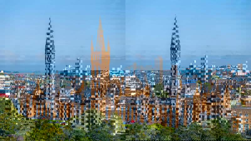 A panoramic shot of the historic Glasgow University building with its striking Gothic revival architecture and prominent clock tower, set against a blue sky with the city skyline in the distance.