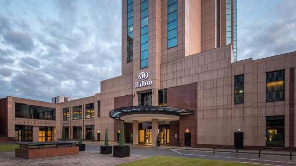 Exterior view of the Hilton Glasgow hotel showing the tall tower, glass entrance canopy, and landscaped forecourt at dusk.