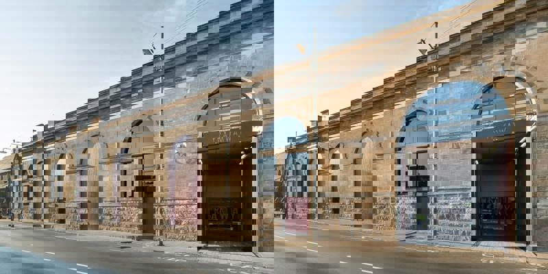 A street view of a large, long, pale stone building with large, industrial arched doorways and windows, with "TRAMWAY" visible above a wide, open entrance.