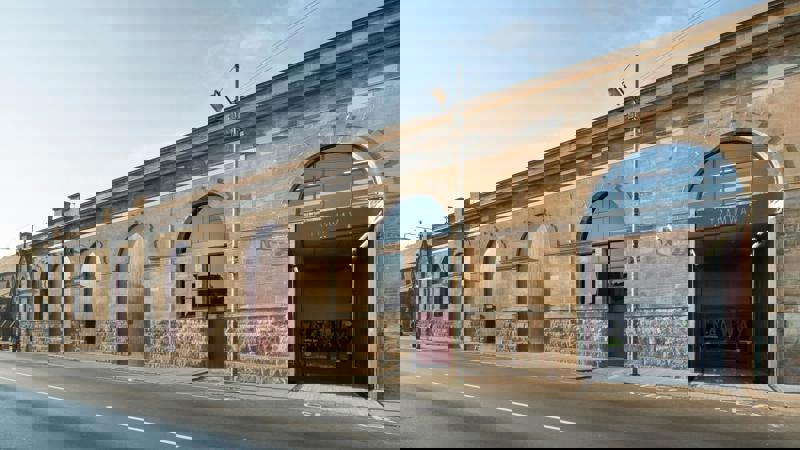 A street view of a large, long, pale stone building with large, industrial arched doorways and windows, with "TRAMWAY" visible above a wide, open entrance.