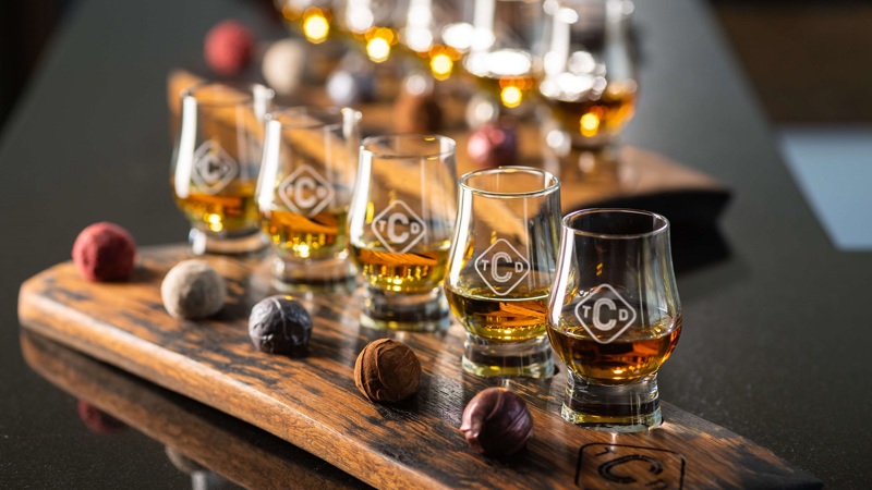 A wooden tasting tray with several glasses of whisky and artisan chocolates.