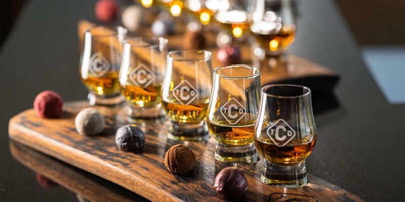 A wooden tasting tray with several glasses of whisky and artisan chocolates.