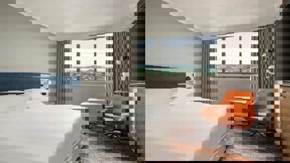 Contemporary hotel room at the Glasgow Marriott featuring a king-size bed, large window with city views, and modern furnishings in neutral tones with an orange accent chair.