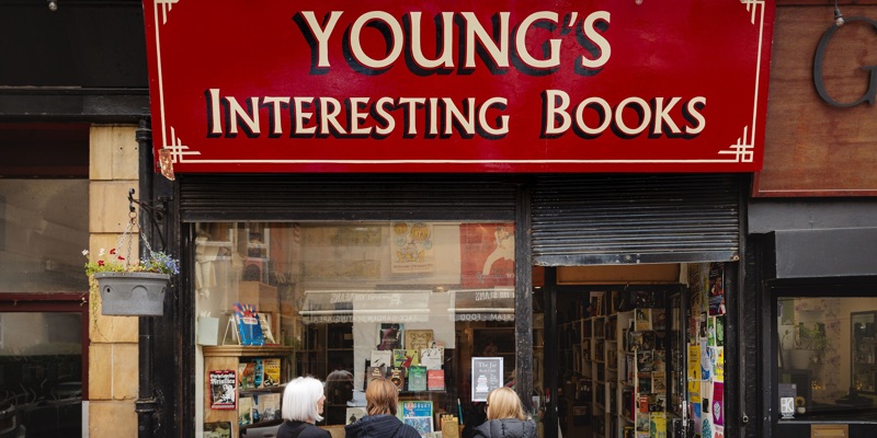 Exterior shot of Young's Interesting Books, a shop with a large red sign and a window display of books. Three people are standing near the entrance, looking inside.