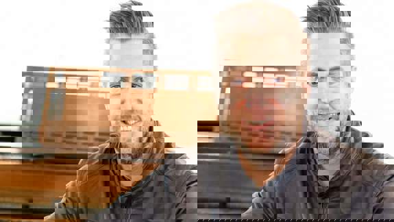 A smiling man in a black leather jacket sitting at a piano with a set of bagpipes on his lap.