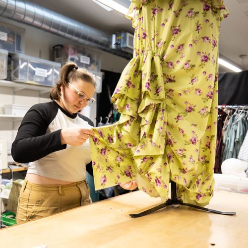 A student is using scissors to cut fabric from a yellow, floral-patterned dress on a table in a workspace filled with sewing supplies and clothing racks.