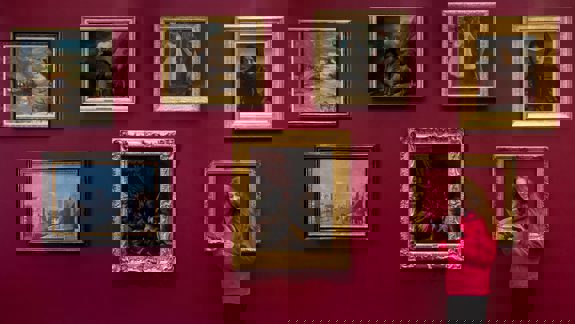 A person in a pink hoodie stands by a red gallery wall with seven framed paintings, looking at a leaflet.