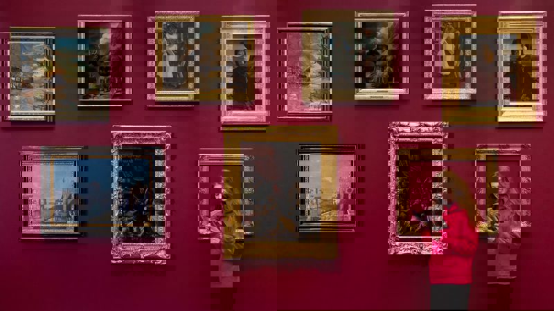 A person in a pink hoodie stands by a red gallery wall with seven framed paintings, looking at a leaflet.