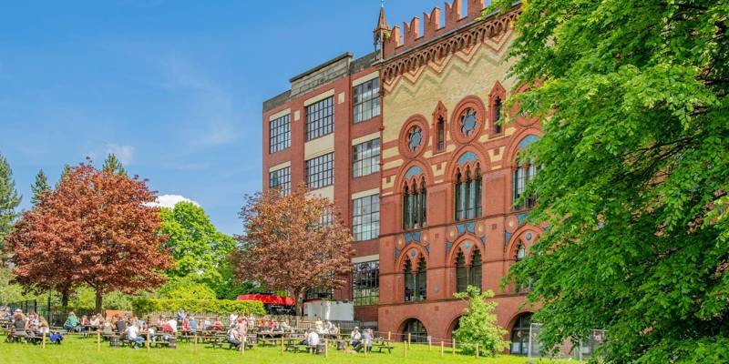 A large, ornate brick building next to a grassy area with people sitting at picnic tables under a sunny sky.